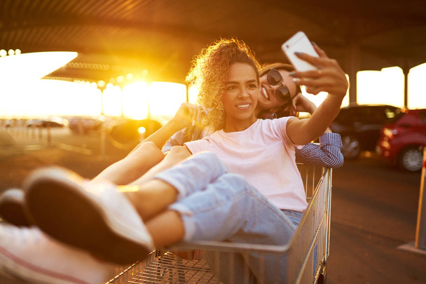 Selfie time! Two beautiful girls having fun on shopping trolleys. Multiethnic young people racing on shopping cart. Beautiful summer day with sunlight. Lifestyle concept. - Foto, imagen