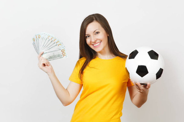 European young fun woman, football fan or player in yellow uniform holding bunch of money banknotes, soccer ball isolated on white background. Sport, play football game, excitement lifestyle concept. - Foto, afbeelding