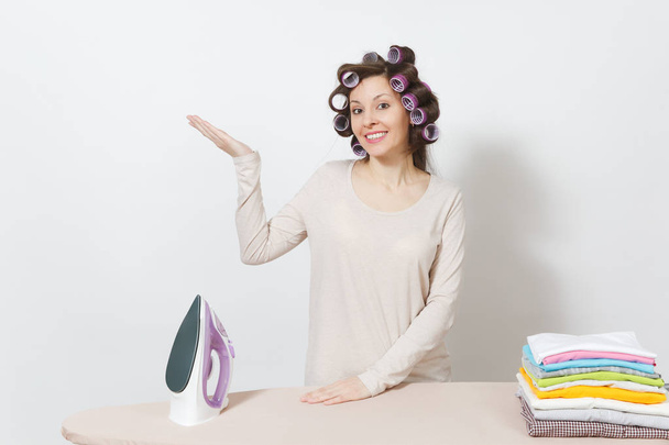 Housewife with curlers on hair in light clothes stand at ironing board with iron, family clothing . Woman point hand aside isolated on white background. Housekeeping concept. Copy space advertisement. - Photo, Image