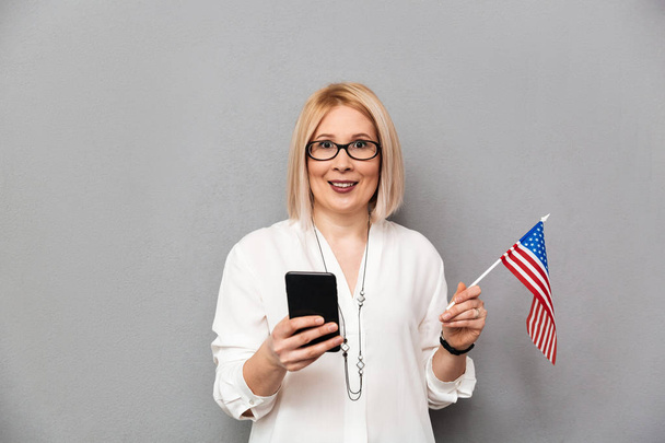 Happy middle-aged woman in shirt and eyeglasses holding USA flag - 写真・画像