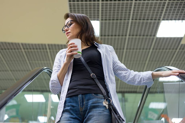 Middle-aged woman with cup of coffee, background shopping mall entertainment center. - Photo, Image
