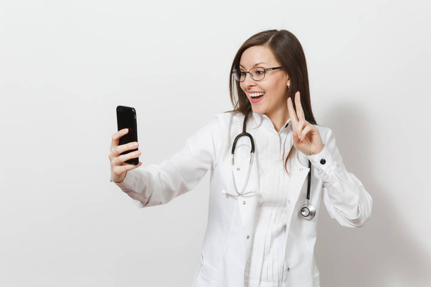 Glimlachend leuke mooie jonge dokter vrouw met stethoscoop, glazen geïsoleerd op een witte achtergrond. Vrouwelijke arts in medische jurk selfie op mobiele telefoon doen. Medisch personeel, gezondheid, geneeskunde conc - Foto, afbeelding