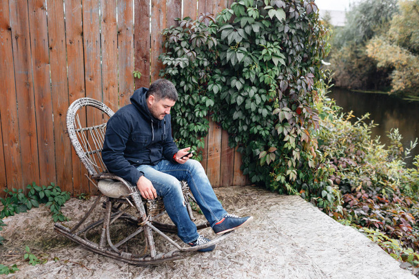 Handsome man relax in vintage rocking-chair with phone in garden. - Zdjęcie, obraz