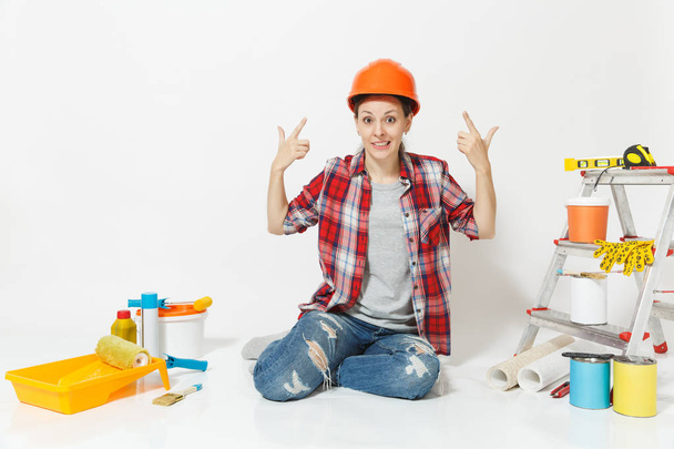 器具を白い背景に分離されたリノベーション アパートの床に座って保護用のオレンジ色のヘルメットで楽しい女。壁紙、接着剤、ペイント ツールのためのアクセサリー。修理の概念. - 写真・画像
