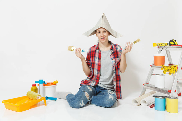 Fun woman in newspaper hat sitting on floor with brush, instruments for renovation apartment room isolated on white background. Wallpaper, accessories for gluing, painting tools. Repair home concept. - Fotografie, Obrázek