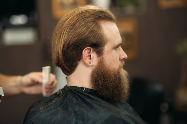 bärtiger Mann, der sich von einem professionellen Friseur mit Kamm und Pflegeschere frisieren lässt. Nahaufnahme mit geringer Schärfentiefe - Foto, Bild