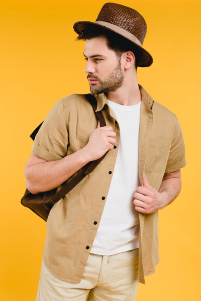 handsome bearded young man in hat holding backpack and looking away isolated on yellow  - Foto, Imagem