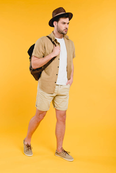 vista completa del joven en pantalones cortos y sombrero sosteniendo la mochila y caminando aislado en amarillo
 - Foto, Imagen