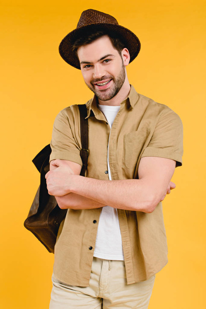 pohledný mladý muž v klobouku stojící s překřížením rukou a usmívá se na kameru izolované na žluté - Fotografie, Obrázek