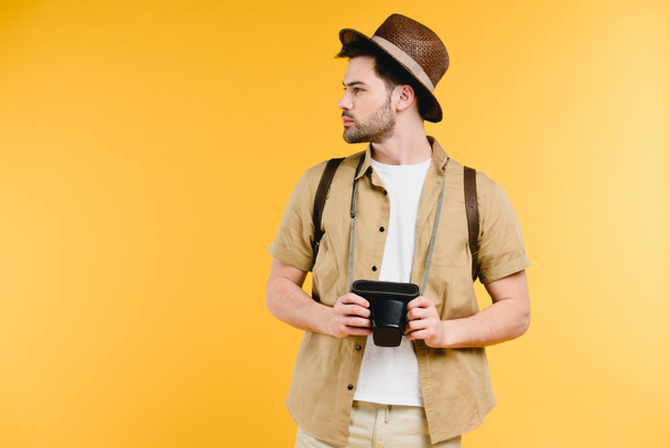 young man in hat with backpack holding camera and looking away isolated on yellow - Fotografie, Obrázek