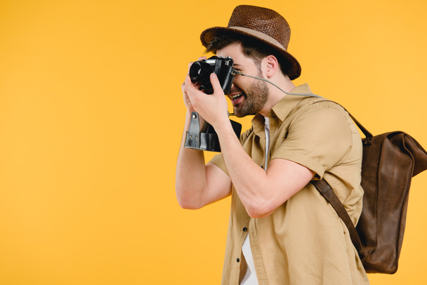 side view of young man in hat with backpack photographing with camera isolated on yellow - Fotó, kép