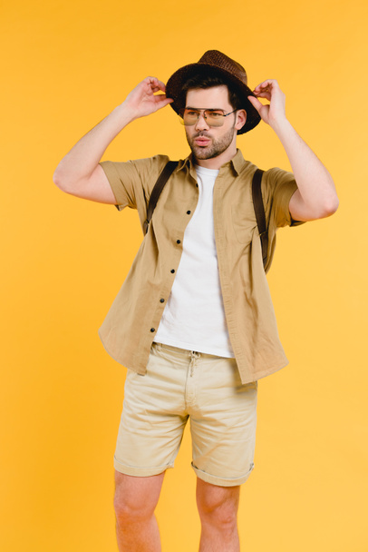 portrait of handsome young man in sunglasses and shorts adjusting hat isolated on yellow  - Photo, Image