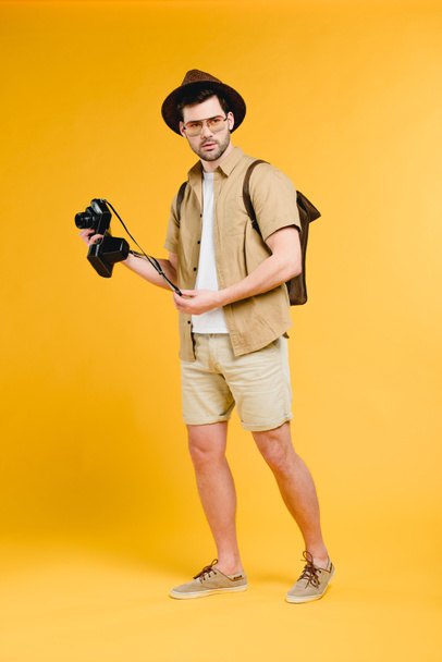 handsome young traveler with backpack holding camera isolated on yellow - Foto, immagini