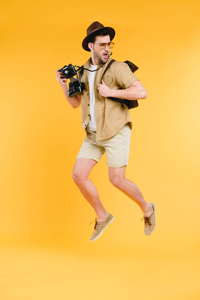 handsome young man in hat and sunglasses holding camera and jumping isolated on yellow - Foto, imagen