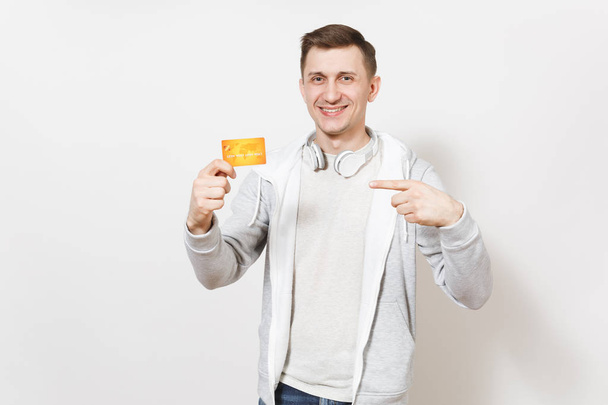 Joven estudiante guapo sonriente en camiseta y sudadera ligera con auriculares alrededor del cuello sostiene la tarjeta de crédito naranja y señala con el dedo en el estudio sobre fondo blanco. Concepto de éxito
 - Foto, Imagen
