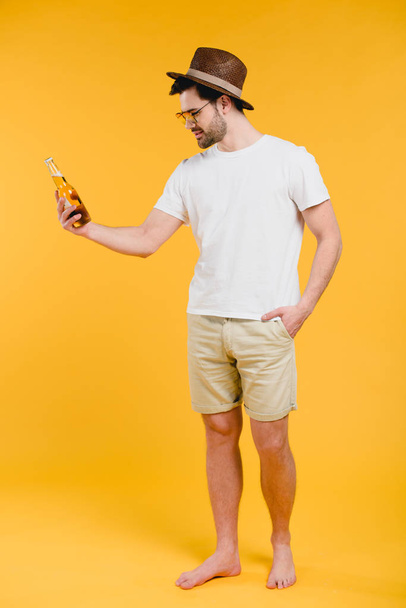 young barefoot man in shorts and hat holding glass bottle with summer drink isolated on yellow - Fotoğraf, Görsel