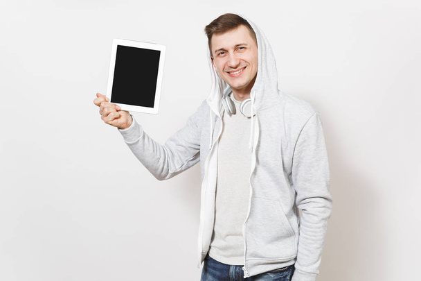 Young handsome smiling student in t-shirt and light sweatshirt with hood with headphones holds a tablet in hands and shows it to camera in studio on white background. Concept of technology - Foto, Imagen