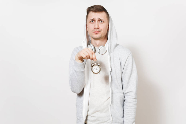 Young handsome embarrassed student in t-shirt and light sweatshirt with hood with headphones holds small alarm clock and shows it in studio on white background. Concept of pastime. Time is running out - Photo, Image