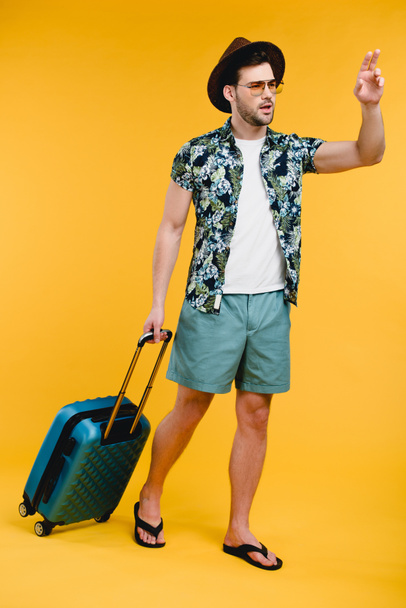 jonge man met koffer op zoek weg en zwaaien hand geïsoleerd op geel - Foto, afbeelding