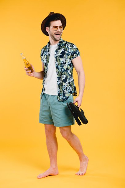 smiling young barefoot man in shorts holding bottle of summer drink and flip-flops isolated on yellow - Photo, Image