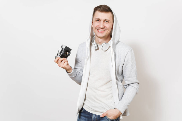 Νεαρό όμορφος χαμογελώντας φοιτητή σε t-shirt και ελαφρύ φούτερ με κουκούλα με ακουστικά δείχνει κάμερας ρετρό και κατέχει το χέρι στην τσέπη στο studio σε λευκό φόντο. Έννοια της φωτογραφίας, χόμπι - Φωτογραφία, εικόνα
