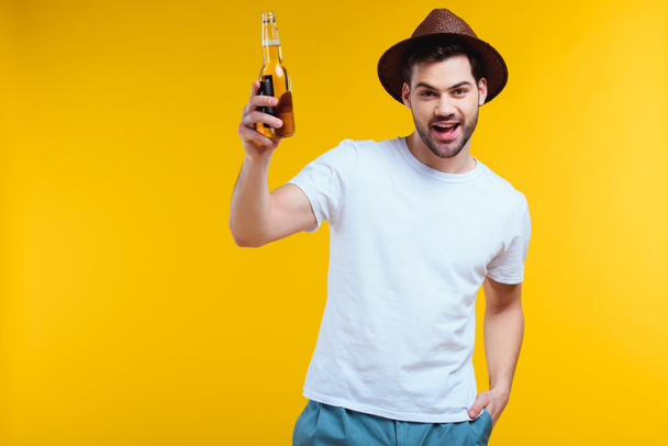 vrolijke jongeman in hoed bedrijf glazen fles van zomer drank en lachend op camera geïsoleerd op geel  - Foto, afbeelding