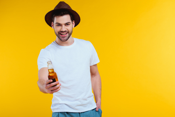 handsome young man in hat holding glass bottle of summer drink and smiling at camera isolated on yellow  - Photo, Image
