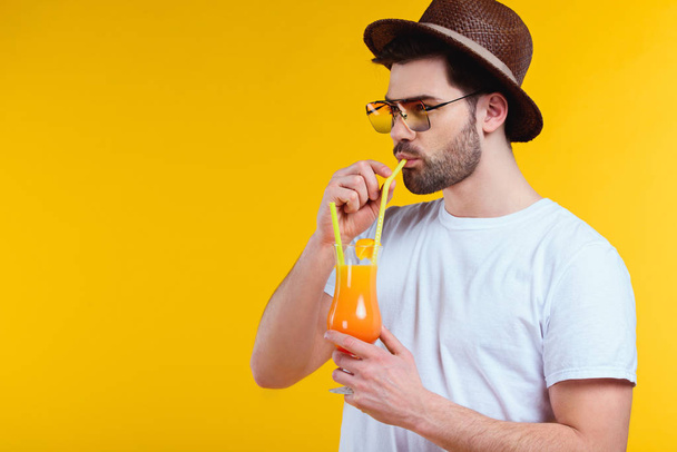 красивий бородатий молодий чоловік у капелюсі та сонцезахисних окулярах п'є літній коктейль ізольовано на жовтому
 - Фото, зображення