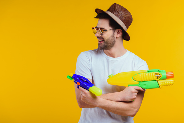 hymyilevä nuori mies hattu ja aurinkolasit leikkii vesi aseet eristetty keltainen
 - Valokuva, kuva