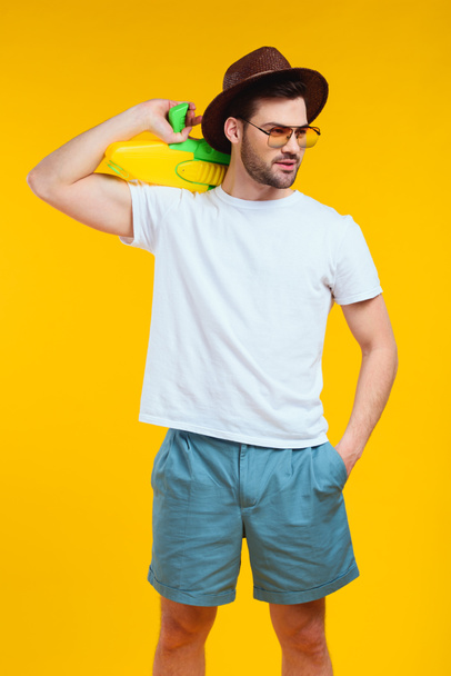 knappe jongeman in zomer outfit houden waterpistool en op zoek weg geïsoleerd op geel met baard - Foto, afbeelding
