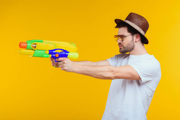 side view of young man holding water gun and looking away isolated on yellow - Photo, Image