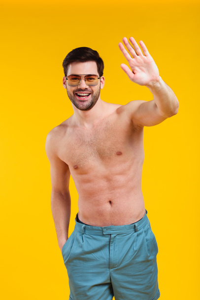 csinos, félmeztelen fiatal férfi nadrág és napszemüveg integetett kézzel és mosolyogva kamera elszigetelt, a sárga - Fotó, kép