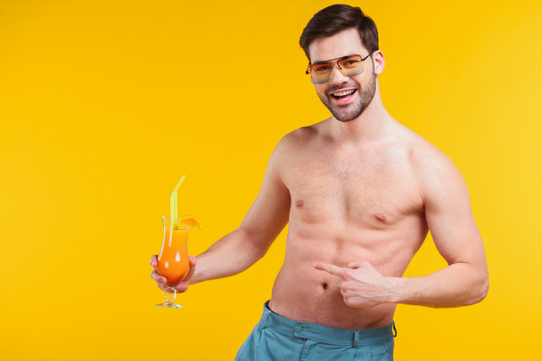 красивый без рубашки молодой человек в шортах держа стакан летнего коктейля и указывая пальцем изолирован на желтый
 - Фото, изображение