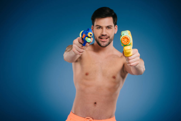 handsome bearded young man holding water guns and smiling at camera on blue  - Photo, Image