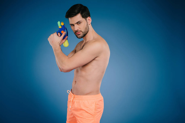 handsome young man in shorts holding water gun and looking at camera on blue - Photo, image