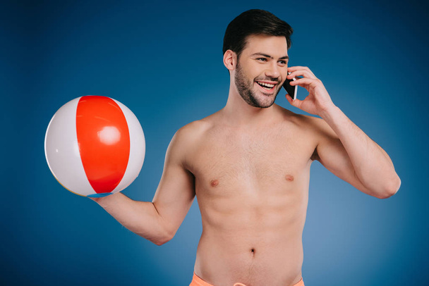 uśmiechający się młody człowiek shirtless trzymając piłkę plażową i rozmawiać przez smartfon na niebiesko - Zdjęcie, obraz