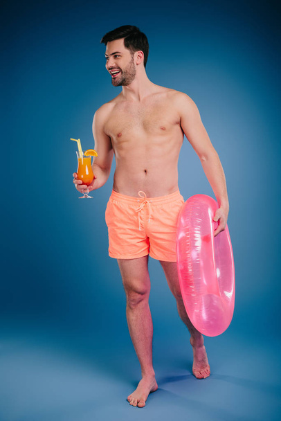 smiling young man in shorts holding glass of summer cocktail and inflatable ring on blue - Fotografie, Obrázek