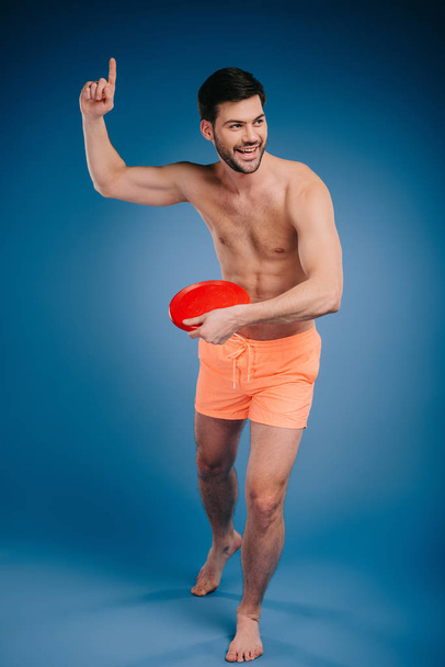 full length view of smiling young barefoot man in shorts holding flying disk and looking away on blue - Foto, Imagen