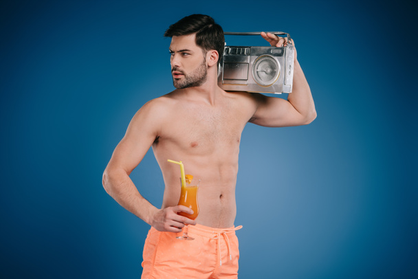 schöner junger Mann in kurzen Hosen mit Tonbandgerät und einem Glas Sommercocktail auf blauem Grund  - Foto, Bild