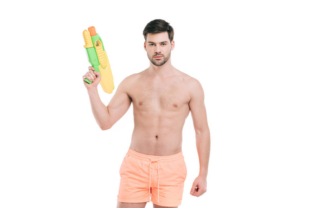 Красивый молодой человек в шортах, держащий водяной пистолет и смотрящий на камеру, изолированную на белом
 - Фото, изображение