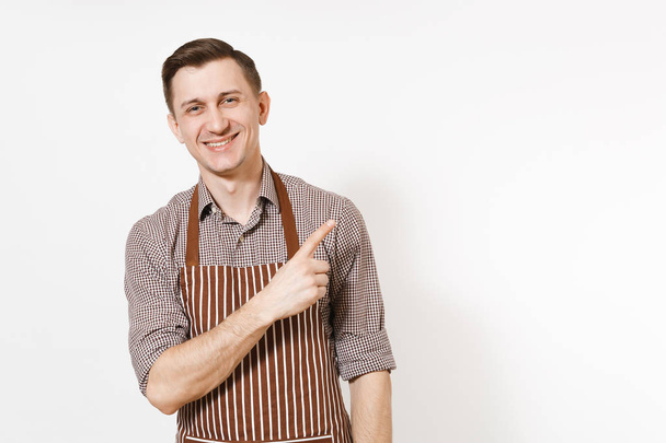Nuori hymyilevä mies kokki tai tarjoilija raidallinen ruskea esiliina, paita osoittaa sormella syrjään kopiotilaa eristetty valkoisella taustalla. Miestaloudenhoitaja tai taloudenhoitaja. Kotimainen työntekijä mainontaa varten
. - Valokuva, kuva