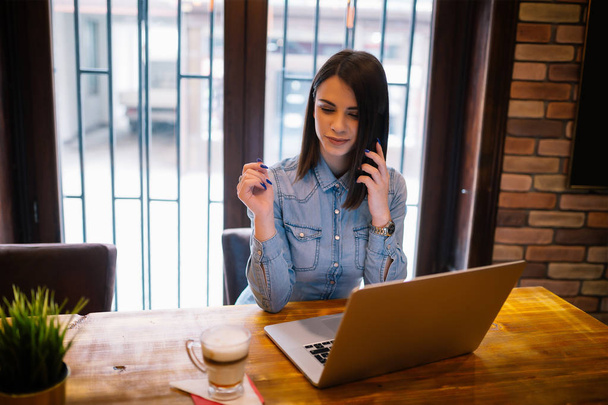 Mladá žena sedí v kavárně u dřevěného stolu, pití kávy a povídali na smartphone. Na stole je notebook. Dívka, procházení Internetu, chatování, blogování. - Fotografie, Obrázek
