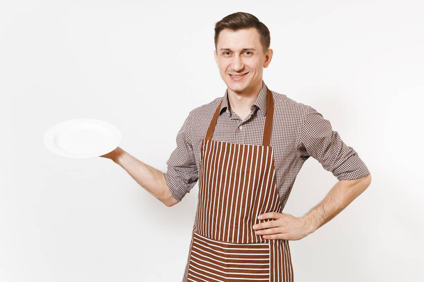 Young man chef or waiter in striped brown apron, shirt holding white round empty clear plate isolated on white background. Male housekeeper or houseworker. Domestic worker copy space for advertisement - Photo, Image