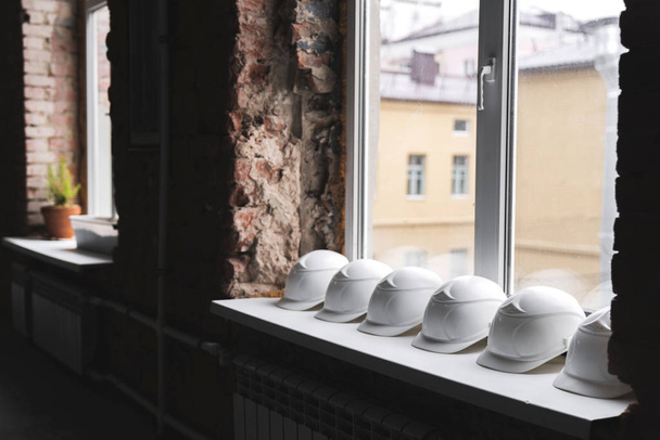 Valkoinen rakentaminen kypärät makaavat ikkunalaudalla peräkkäin rakennuksen sisällä rakenteilla. rakennus- ja ryhmätyön käsite
. - Valokuva, kuva