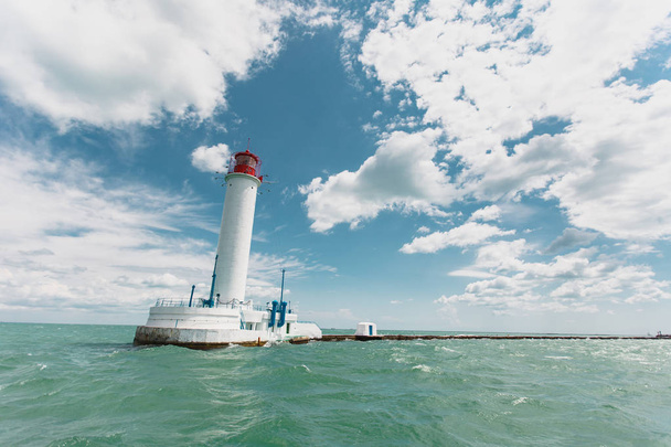 Hermoso paisaje marino. Un faro de torre guía de identificación costera blanca alta con una cabecera roja destinada a la navegación de barcos en medio del mar azul ondulado contra un cielo azul nublado
 - Foto, imagen