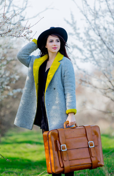 Young redhead girl in coat and hat with suitcase on springtime outdoor  - Фото, зображення