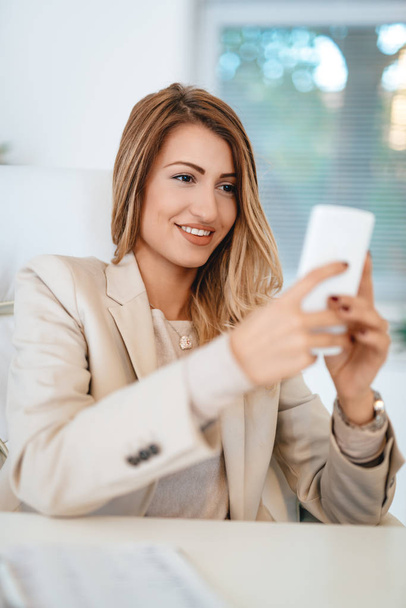 Молодая симпатичная улыбающаяся деловая женщина пишет смс на смартфоне в офисе
.  - Фото, изображение