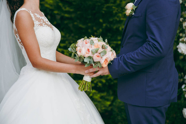 Krásné svatební oslavu. Ženich v modrém obleku s boutonniere a nevěsta v elegantní krajkové šaty se závojem a kytice pivoněk a zelené listy, drželi se za ruce v zelené zahradě - Fotografie, Obrázek