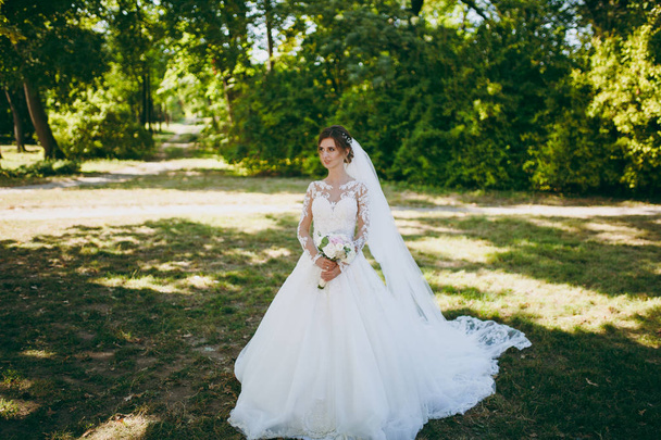 Hermosa sesión de fotos de boda. La novia en un vestido de encaje blanco con una pluma larga, horquilla en el pelo, velo y ramo de flores blancas y rosadas en un gran jardín verde en el día soleado y lluvioso
 - Foto, Imagen