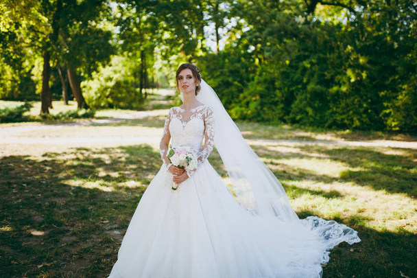 Прекрасная свадебная фотосессия. Невеста в белом кружевном платье с длинным шлейфом, шпилька в волосах, вуаль и букет белых и розовых цветов в большом зеленом саду в солнечный день
 - Фото, изображение